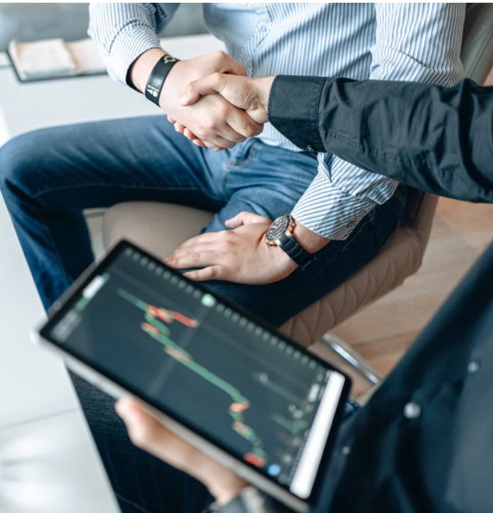 Two executives shaking hands over a tablet showing financial charts